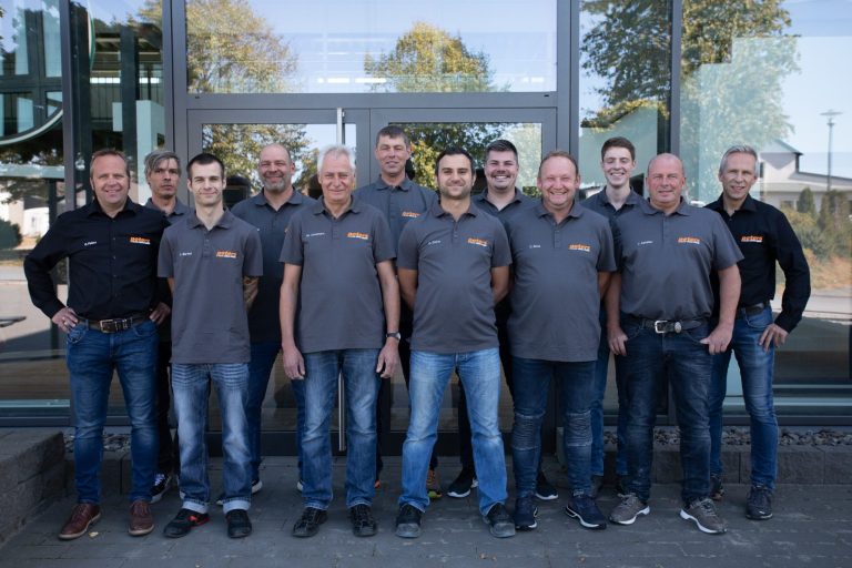 Gruppe von 15 Personen in grauen T-Shirts, stehend vor einem modernen Gebäude.