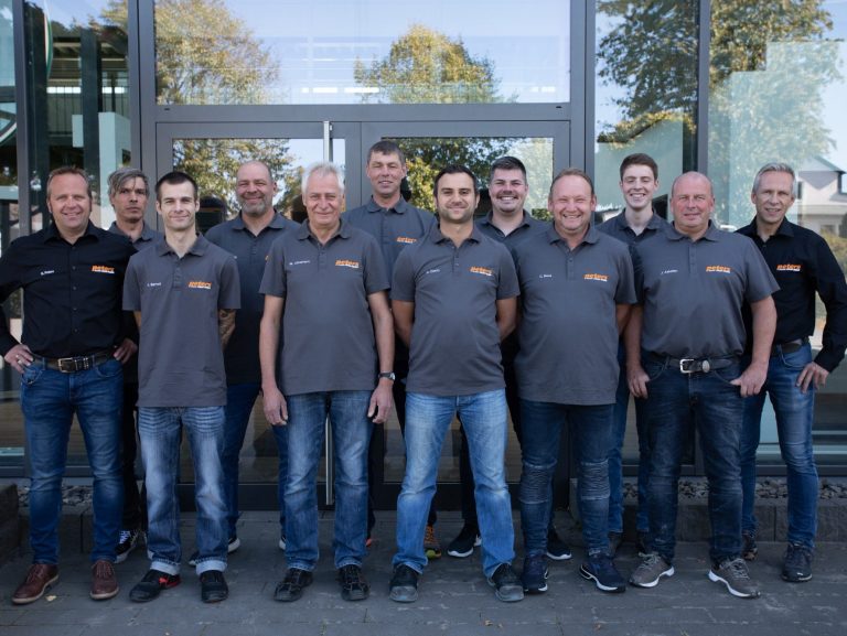 Gruppfoto von zehn Mitarbeitern der Firma Peters in grauen T-Shirts mit dem Firmenlogo, die lächeln und vor einer Glasfassade stehen.