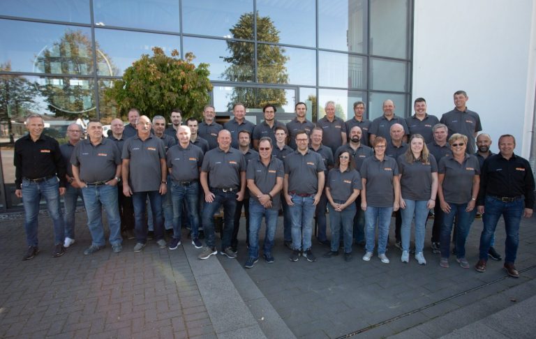 Mitarbeiterfoto von 37 Personen in grauen Shirts mit Peters Logo vor einem modernen Gebäude.