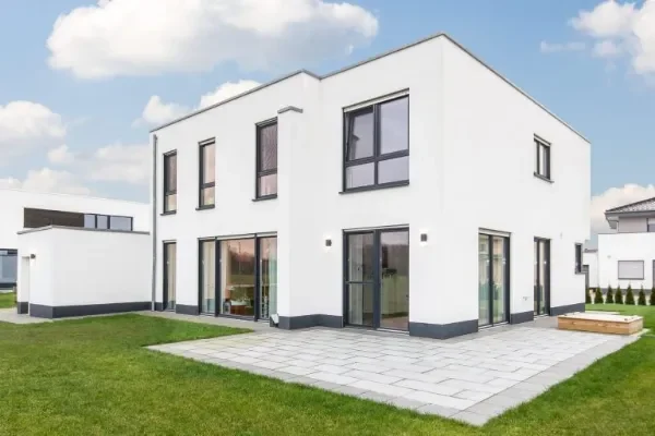 Modernes, weißes Einfamilienhaus mit großen Fenstern und Terrasse in einem grünen Umfeld.