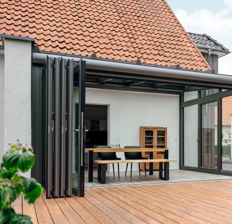 Moderne Terrasse mit Holzdecke, Glaswänden und einem Esstisch aus Holz und Stühlen.