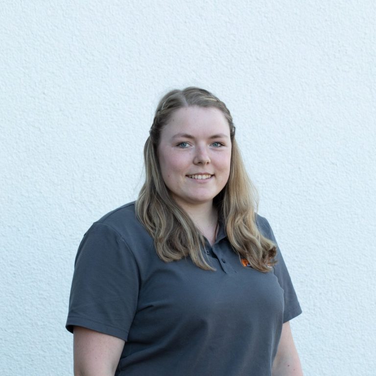 Junge Frau mit langen, blonden Haaren, lächelt in grauem Poloshirt vor hellem Hintergrund.