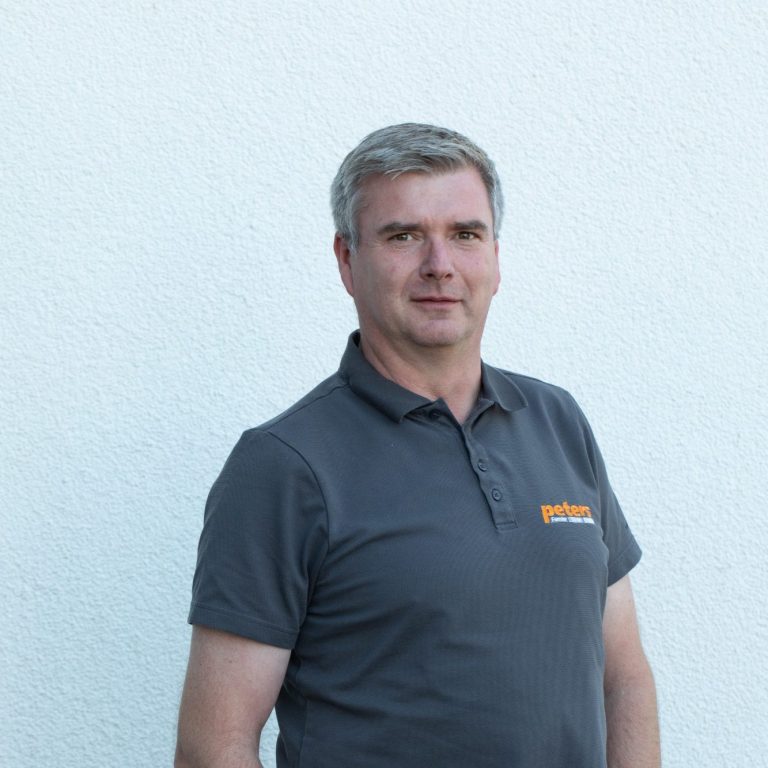 Mann mit grauem Shirt steht vor einer hellen Wand.