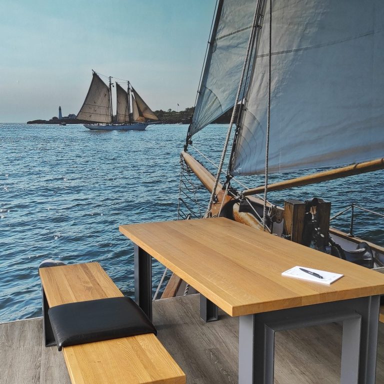 Holztisch mit Bank vor maritimer Wandgestaltung und Segelschiff auf dem Wasser.