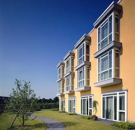 Moderne, gelbe Gebäude mit großen Fenstern und grünem Garten bei blauem Himmel.
