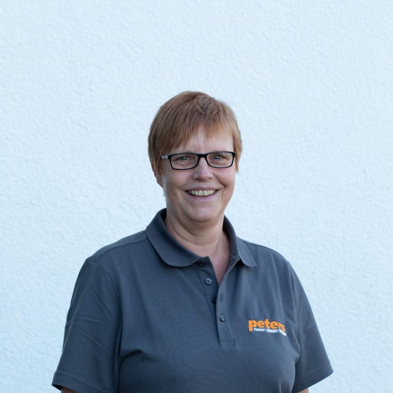 Lächelnde Frau mit Brille in grauem Polo-Shirt vor hellem Hintergrund.