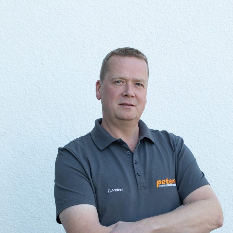 Mann in grauem Poloshirt mit verschränkten Armen, stehend vor weißer Wand.