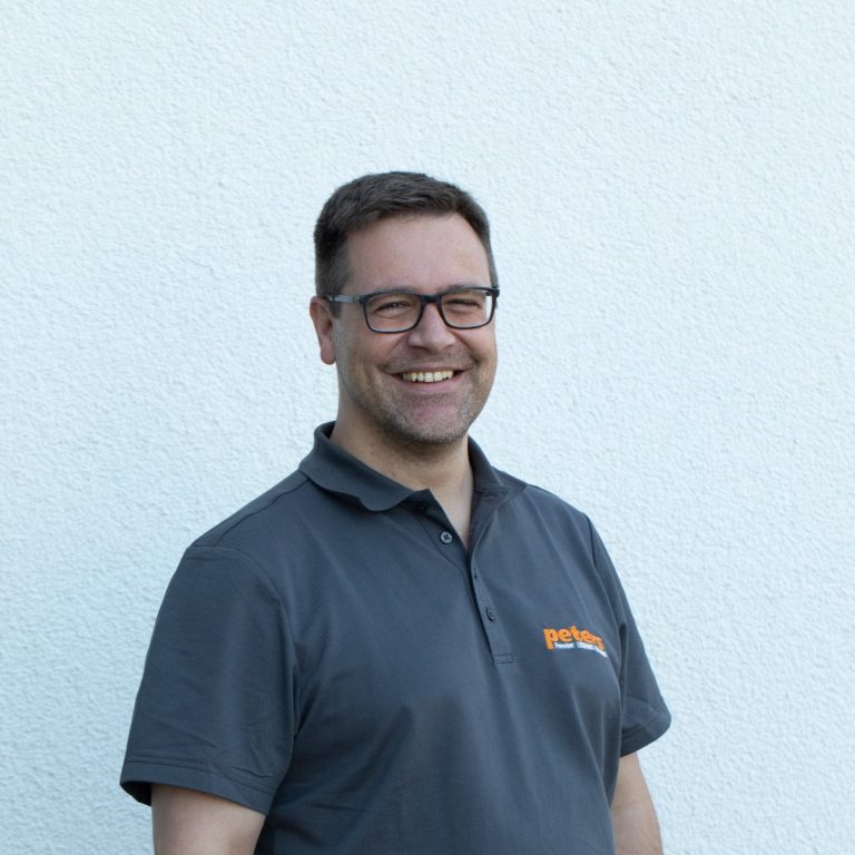 Mann mit Brille, lächelnd, trägt ein graues Polo-Shirt vor hellem Hintergrund.