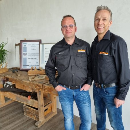 die zwei Geschäftsführer in schwarzen Hemden stehen neben einet Werkbank aus Holz mit Urkunden und Werkzeug im Hintergrund.