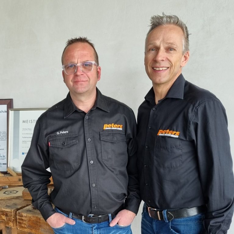 Zwei Männer in schwarzen Hemden stehen lächelnd nebeneinander vor Zertifikaten.