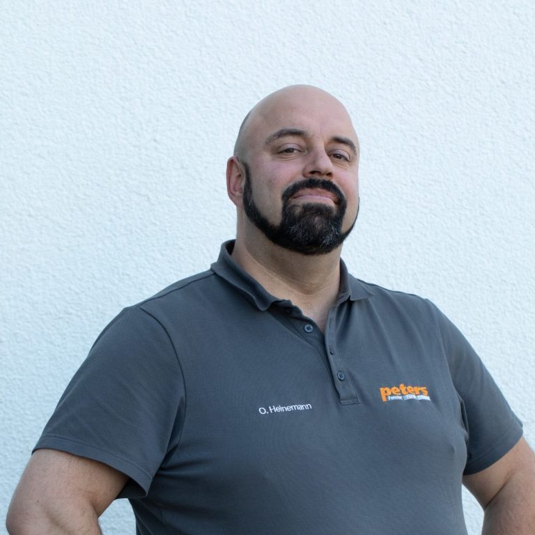 Mann mit Bart steht lächelnd vor einer hellen Wand, trägt ein graues Polohemd.